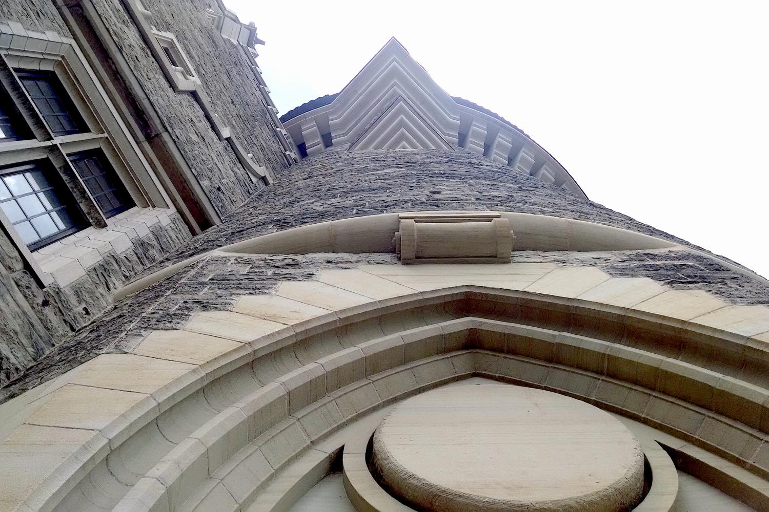 Upward view of Casa Loma’s stone tower and Gothic Revival facade, featuring arched doorway, varied masonry textures, wood, and multi-pane windows.